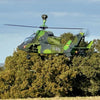 Model Green military helicopter flying over trees with a clear blue sky.